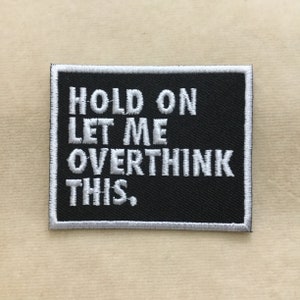 May include: Black rectangular patch with white embroidered text that reads "HOLD ON LET ME OVERTHINK THIS." The patch has a white border and is set against a neutral background.
