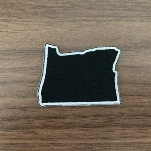 May include: An embroidered patch in the shape of the state of Oregon. The patch features a solid black center with a thick, white stitched border outlining the entire perimeter of the state's geographic shape, creating a high-contrast design.