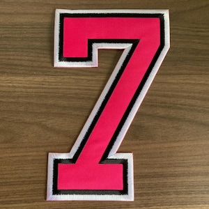 May include: A large, fabric number seven patch featuring a vibrant hot pink center. The number is outlined with a thick black border, followed by a thin white stitched edge that defines the shape against the background surface.