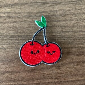 May include: Embroidered patch featuring two smiling red cherries with black stems and green leaves. The cherries have black outlines and white stitching. The patch is on a wooden surface.