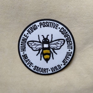 May include: Round embroidered patch featuring a stylized bee with black and yellow stripes and wings. The patch has a white background with the words "HUMBLE, KIND, POSITIVE, CONFIDENT, JOYFUL, WILD, SMART, BRAVE" in a black border.