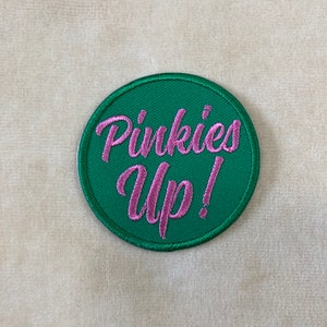 May include: A round, green embroidered patch with the phrase "Pinkies Up!" in pink script. The patch has a stitched border and is set against a neutral, textured background.
