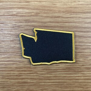 May include: A black embroidered patch in the shape of the state of Washington, with a gold border. The patch is on a wooden surface.