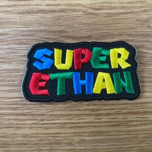 May include: A colorful embroidered patch with the words "SUPER ETHAN" in a playful, blocky font. The text is in blue, yellow, red, green, and black, with a black border. The patch is likely for clothing or accessories.