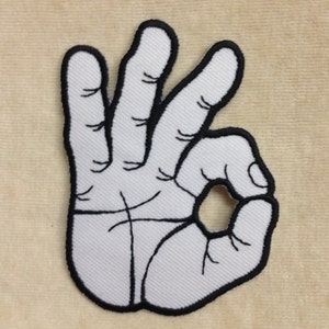 May include: White embroidered patch in the shape of a hand making the "OK" gesture. The patch has a black outline and detailed stitching. The background is a light beige fabric.