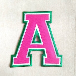 May include: Embroidered letter "A" patch in a collegiate style. The letter is a vibrant pink with a green border and white accents. The patch is likely for jackets, bags, or other fabric items.