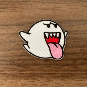 May include: An embroidered patch featuring a white ghost character with a black outline, sharp black eyebrows, a wide open mouth with white teeth, a red interior, and a long pink tongue sticking out, all rendered in a cartoon style.