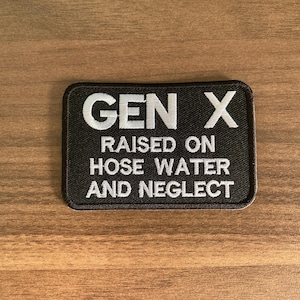 May include: Black rectangular patch with white embroidered text that reads "GEN X RAISED ON HOSE WATER AND NEGLECT". The patch is on a wooden surface.