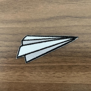 May include: A white and black embroidered paper airplane patch. The patch features a detailed design with black outlines and shading on a white background. The patch is on a wooden surface.