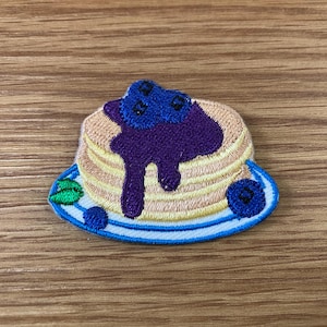 May include: Embroidered patch featuring a stack of pancakes with blueberry syrup and blueberries on a blue plate. The patch has a detailed, stitched design with tan, purple, blue, and green colors. The patch is on a wooden surface.