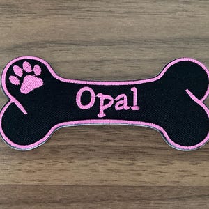 May include: A bone-shaped embroidered patch featuring a black background with a bright pink border. The patch displays a pink paw print on the left side and the name Opal embroidered in pink lettering across the center of the bone shape.