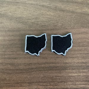 May include: Two black Ohio state-shaped patches with white embroidered borders. The patches are placed on a wooden surface. These patches could be used to decorate clothing or accessories.