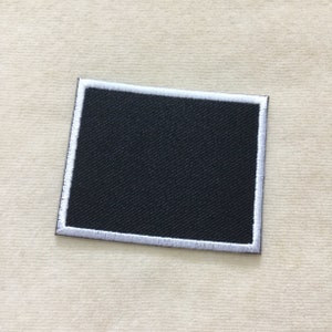 May include: A rectangular black patch with a white embroidered border. The patch has a textured surface and is likely made of fabric. The design is simple and clean, suitable for various applications.