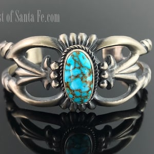 Puede incluir: Brazalete de plata con una piedra de turquesa ovalada en el centro. La turquesa es de un azul vibrante con matriz marrón. El brazalete tiene un diseño calado ornamentado con detalles florales y de volutas. El texto "East of Santa Fe.com" es visible.