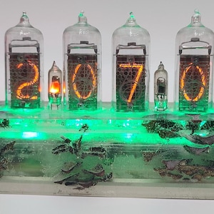 Könnte beinhalten: Eine Digitaluhr mit sechs Glasröhren, die die Uhrzeit 12:07:05 in leuchtendem Orange anzeigen. Die Uhr befindet sich in einem transparenten Harzsockel mit eingebauten grünen LED-Leuchten und dekorativen Elementen.