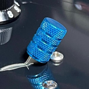 May include: Close-up of a bright blue guitar knob with a textured surface. The knob is cylindrical with horizontal grooves and is attached to a silver metal lever and a silver screw. The background is dark.