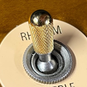 May include: Close-up of a gold-colored knob with a diamond pattern on a guitar. The knob is labeled "Rhythm".