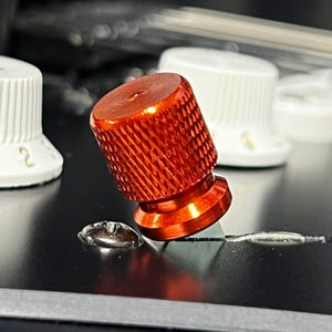 Op de afbeelding: Close-up van een oranje, getextureerde knop met een geribbeld ontwerp, waarschijnlijk voor een muziekinstrument of elektronisch apparaat. De knop staat op een donkere ondergrond, met andere witte knoppen op de achtergrond.