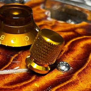 Puede incluir: Primer plano de una perilla de color dorado con textura moleteada, montada sobre una superficie de madera marrón. La perilla está al lado de una perilla más grande, de color dorado, con una parte superior de plástico transparente y los números 8, 9 y 10 visibles.