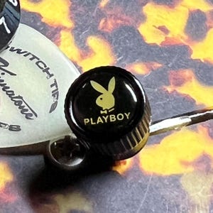 May include: A black cylindrical guitar control knob featuring a gold Playboy rabbit logo and the text PLAYBOY printed on the top surface. The knob has a ribbed edge for grip and is positioned next to a white guitar switch tip component.