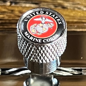 Puede incluir: Un tapón de válvula de metal plateado con un emblema negro y rojo que dice "United States Marine Corps".