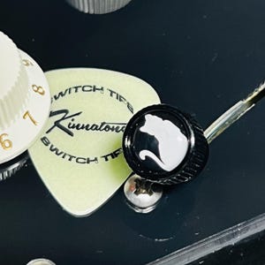 Puede incluir: Un mando de guitarra negro con la silueta de un gato blanco, situado junto a una púa de guitarra de color amarillo pálido con el texto Kinnatone Switch Tips impreso en negro, sobre una superficie negra lisa con un mando numerado blanco.