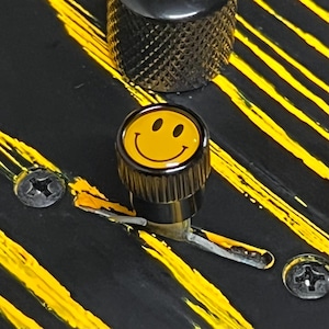 May include: Close-up of a black guitar with yellow stripes and a smiley face knob. The knob is yellow with a black smiley face. Two screws are visible, along with a second black knob.