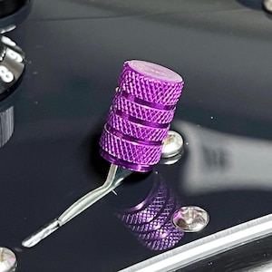 Puede incluir: Primer plano de un botón de guitarra morado con superficie texturizada. El botón es cilíndrico y está unido a un brazo de metal plateado. El fondo es una superficie oscura y brillante con tornillos plateados.