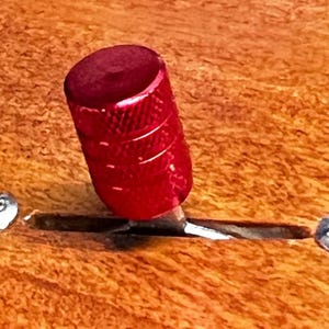 Puede incluir: Primer plano de un botón rojo texturizado con un interruptor de metal plateado. El botón es cilíndrico con una parte superior redondeada y está colocado sobre una superficie de madera. La madera tiene un tono marrón cálido.