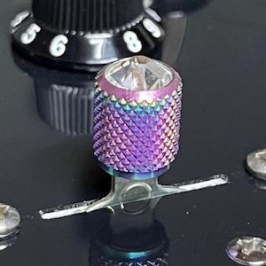 Puede incluir: Primer plano de un botón de guitarra con una parte superior facetada e iridiscente y un cuerpo texturizado púrpura y azul. El botón está montado sobre una superficie negra, con un panel de control negro y números visibles en el fondo.