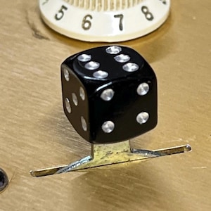 Puede incluir: Un pomo de dados de plástico negro con puntos blancos, montado sobre una base de metal plateado. El pomo está fijado a una superficie de madera.