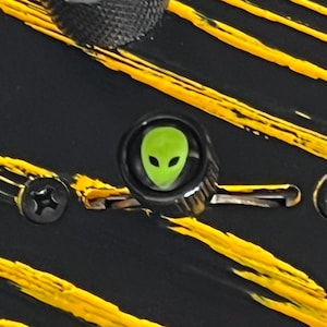 May include: Close-up of a black guitar with yellow paint strokes. A green alien head is centered on a knob. The guitar has a black screw and a textured knob.