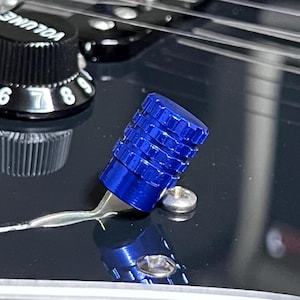 May include: Close-up of a blue guitar tremolo arm tip. The tip is cylindrical with a ridged design. A black volume knob with white numbers is visible in the background. The guitar's body is black.