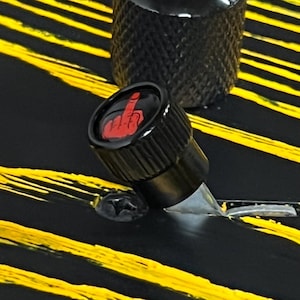 May include: Close-up of a black guitar knob with a red middle finger graphic. The knob is set against a black surface with yellow stripes. Another black knob is visible in the background.