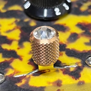 Pode incluir: Um botão de controlo de guitarra cilíndrico em metal dourado com aderência serrilhada em padrão de diamante e uma incrustação de cristal transparente facetado no topo, posicionado sobre um corpo de instrumento com padrão de tartaruga amarelo e preto.