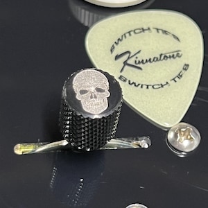 May include: Close-up of a black guitar switch tip with a skull design. A light green guitar pick with the text "Switch Tip" and "Kinnatone" is next to the switch tip. A white guitar knob with numbers is in the background.