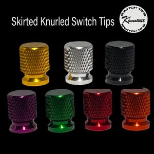 May include: A collection of Skirted Knurled Switch Tips in various colors, including gold, silver, black, purple, green, red, and orange. The tips have a textured, knurled surface and a skirted base. The text "Skirted Knurled Switch Tips" is displayed.