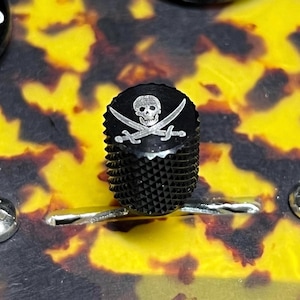 Puede incluir: Un mando de control de guitarra cilíndrico negro con un lateral moleteado texturizado y una superficie superior plana. La parte superior presenta un gráfico grabado en tono plateado de un cráneo humano sobre dos espadas cruzadas, emblema Jolly Roger.