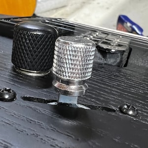 Puede incluir: Primer plano de una perilla plateada con un diseño moleteado en una guitarra negra. La perilla está unida a un eje que se extiende hacia abajo en el cuerpo de la guitarra.