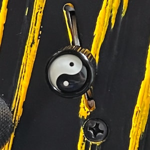 May include: Close-up of a black and white Yin Yang symbol on a black surface. The symbol is centered within a black circular frame, with a silver metal clasp above. Yellow paint streaks are visible in the background.