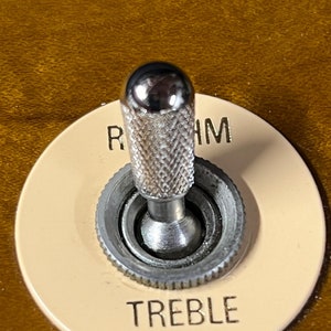 以下が含まれることがあります： クロムメッキのノブのクローズアップ。ノブは、金色の「リズム」と「トレブル」の文字が書かれたベージュのノブに取り付けられています。ノブの表面は、ローレット加工が施されています。