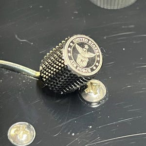Può includere: Una manopola del volume per chitarra in metallo nero con impugnatura zigrinata e un inserto circolare superiore che mostra l'emblema della United States Air Force, con il testo United States Air Force, posizionata su un corpo chitarra nero.