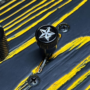 Puede incluir: Primer plano de una guitarra negra con rayas amarillas y un botón en forma de estrella. El cuerpo de la guitarra tiene un acabado envejecido, con tornillos visibles y una superficie texturizada. El botón presenta un diseño de estrella blanca.