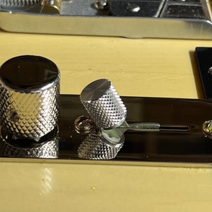 May include: Close-up of a silver guitar control knob with a knurled surface. The knob is attached to a silver metal plate with a black background.