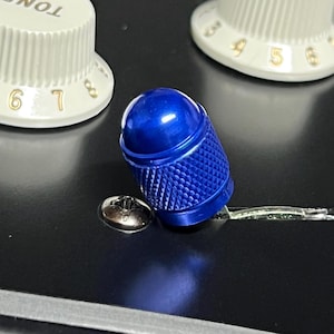 Może przedstawiać: Zbliżenie niebieskiego pokrętła gitary z teksturowanym uchwytem, umieszczonego na czarnej powierzchni. Dwa białe pokrętła z oznaczeniami liczbowymi są widoczne w tle. Widoczna jest również srebrna śruba.