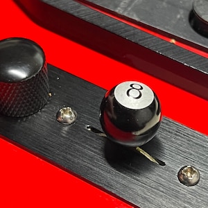 May include: A red electric guitar body featuring a black brushed metal control plate. The plate includes a textured black volume knob, two silver screws, and a black selector switch topped with a spherical black 8-ball knob with a white circle.
