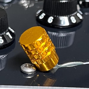 May include: Close-up of a gold-colored, cylindrical knob with a ridged design, likely for an electronic device. The knob sits on a dark surface, near a silver screw and a white wire. Black knobs with white numbers are visible in the background.