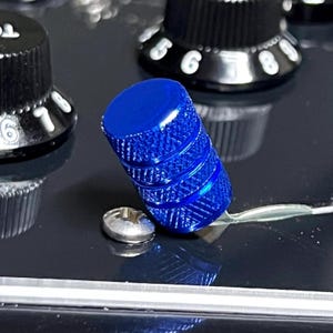 May include: Close-up of a bright blue knob with a ridged texture, positioned next to a silver screw and a black control panel. Other black knobs with white numbers are visible in the background.