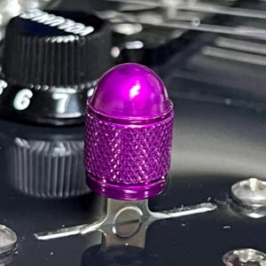 Puede incluir: Primer plano de un botón de guitarra morado con un agarre texturizado, colocado sobre una superficie negra. El botón metálico refleja la luz, con una parte superior redondeada. Un botón de control de volumen negro es visible en el fondo.