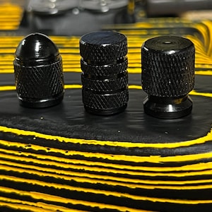 Op de afbeelding: Drie zwarte metalen knoppen met een getextureerd oppervlak worden getoond tegen een zwart-gele achtergrond. De knoppen hebben verschillende vormen, waaronder een kogelvorm, een gestapelde stijl en een afgeronde basis.